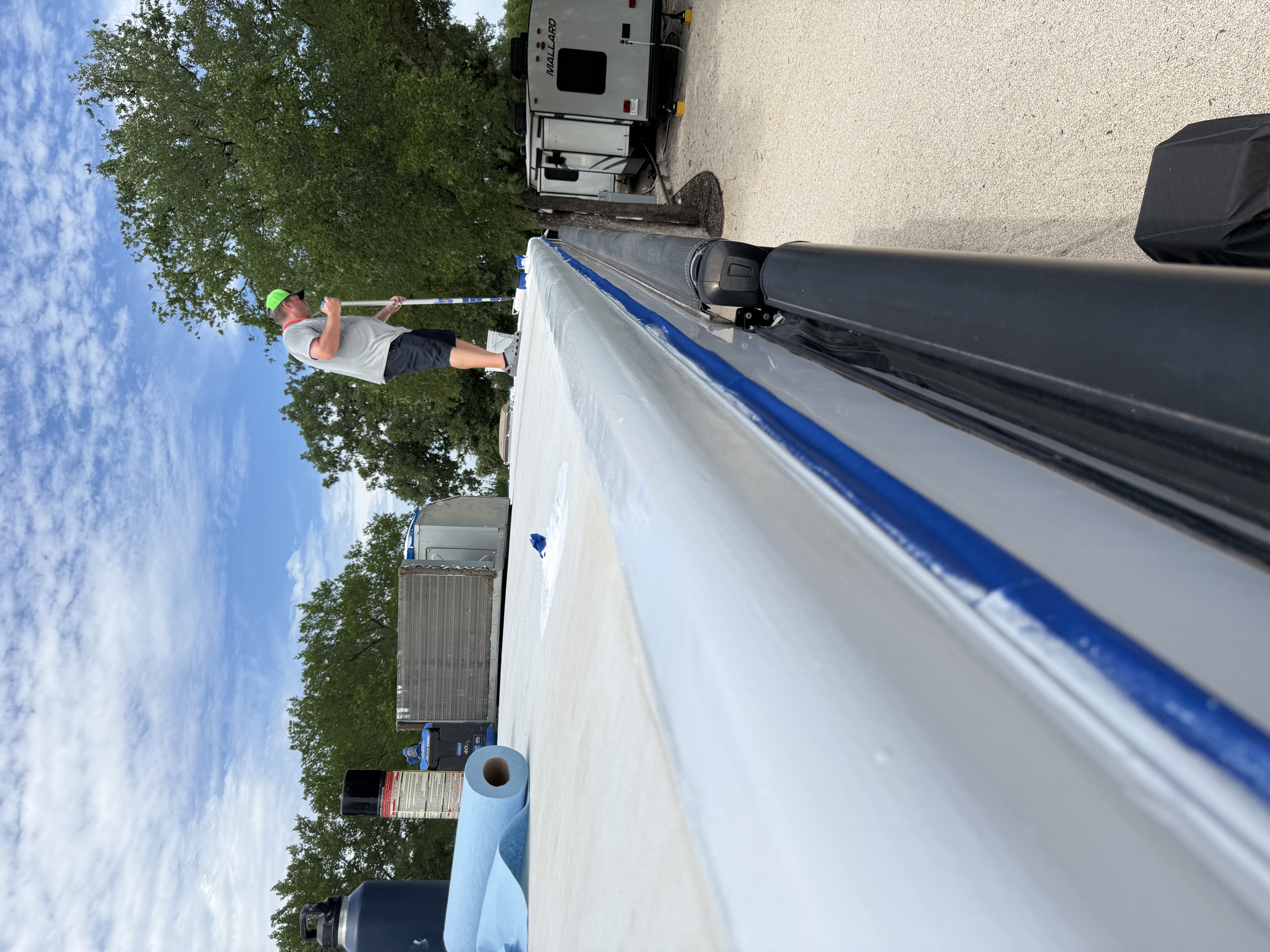 Ground-level view along the roofline of a travel trailer at an RV park — Boss Bull technician standing on the roof applying Crazy Seal with a long-handled roller, blue tape edge seal visible at the drip rail, product cans and blue shop roll on the roof surface — Boss Bull Mobile RV Services