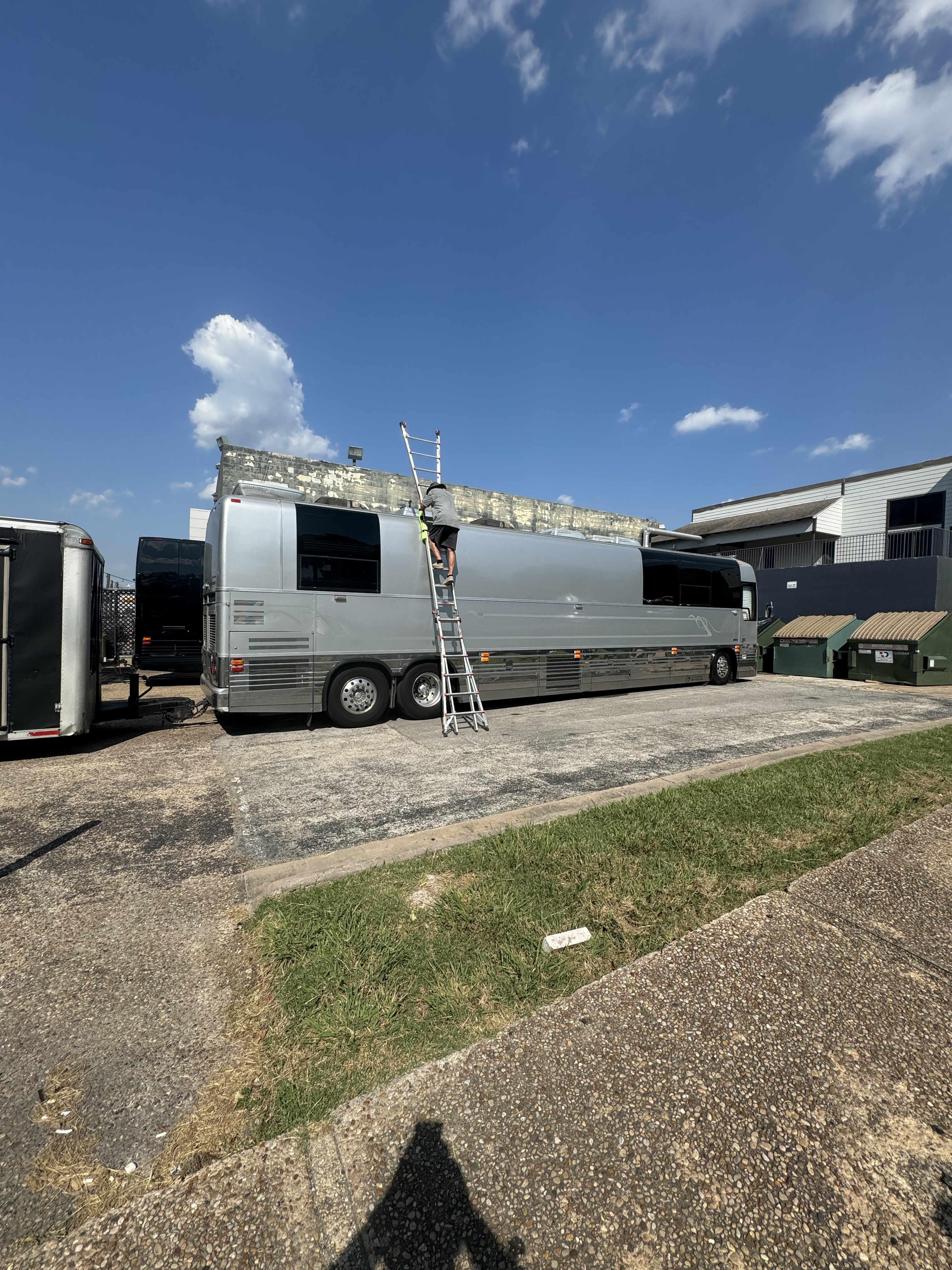 Wide establishing shot of a Boss Bull technician on a ladder mid-climb against the full length of a silver Prevost entertainer coach — mobile roof inspection in progress — Boss Bull Mobile RV Services