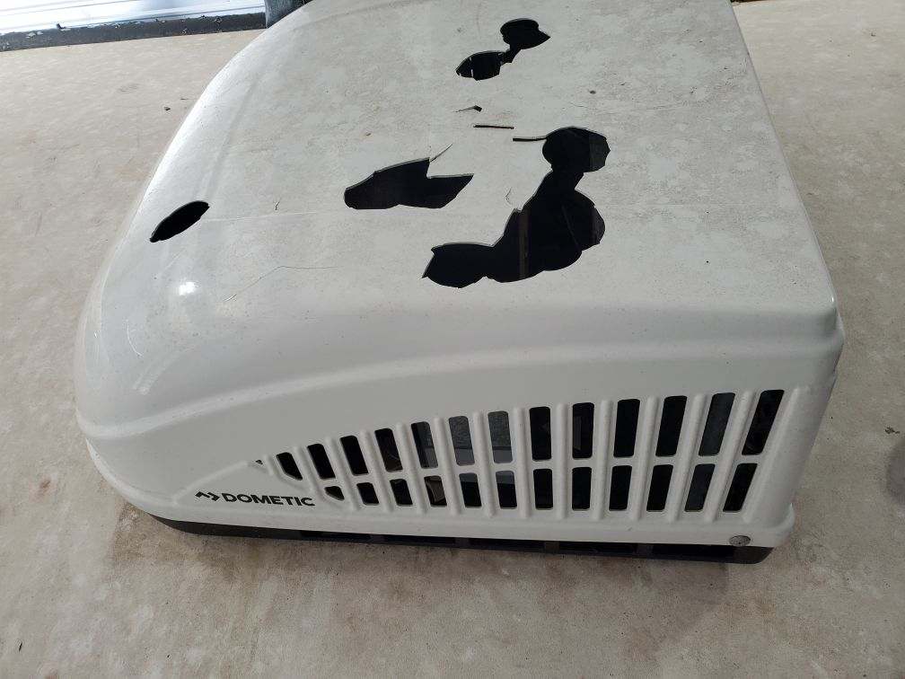 Removed Dometic RV air conditioner shroud on a concrete floor showing catastrophic hail damage — multiple large holes punched through the plastic with jagged broken edges, Dometic logo visible — Boss Bull Mobile RV Services