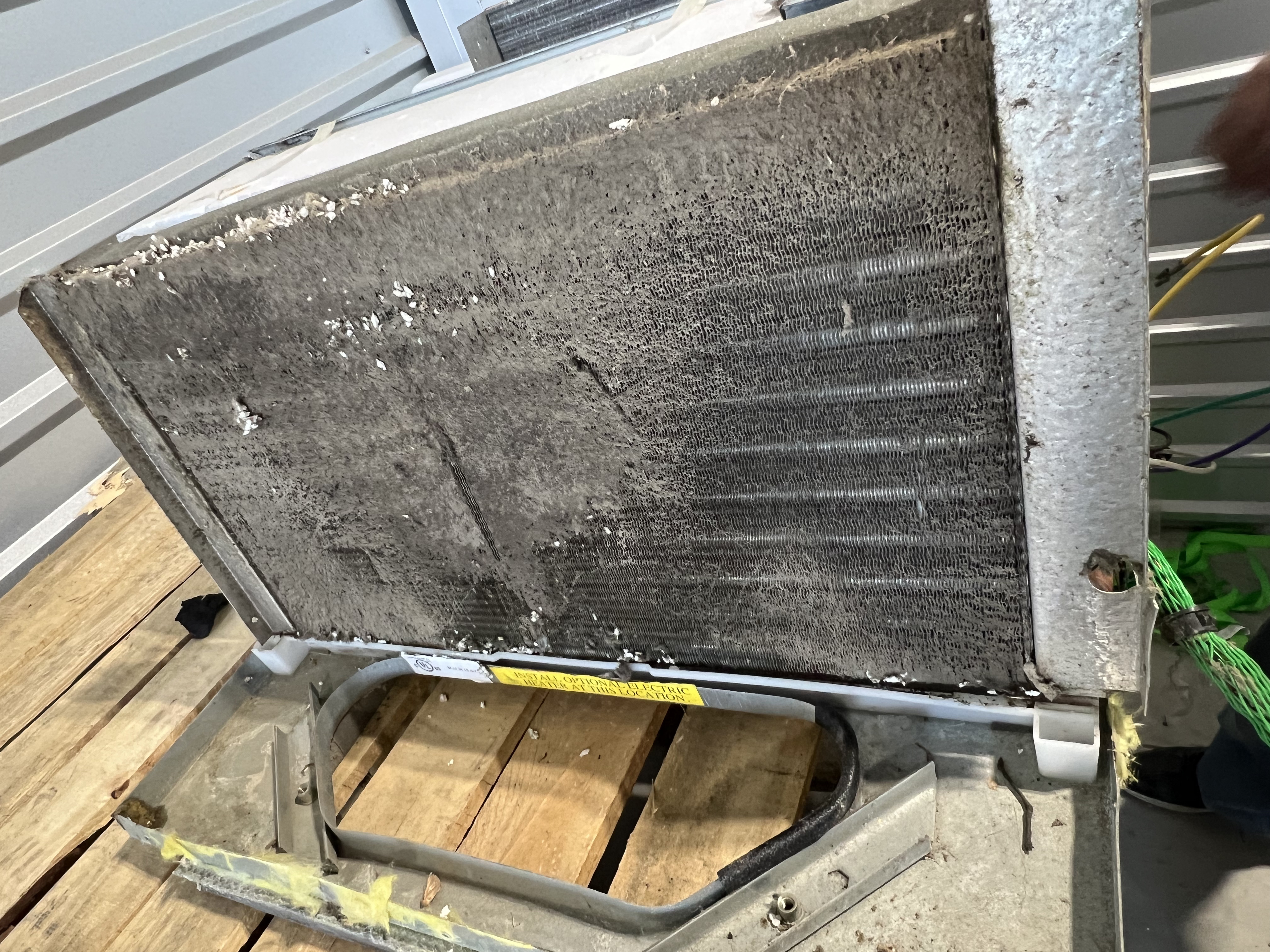 Removed RV AC unit laid flat on a shop floor with the condenser panel propped open at 45 degrees showing the full coil face uniformly packed with dark gray debris across every fin row — Boss Bull Mobile RV Services