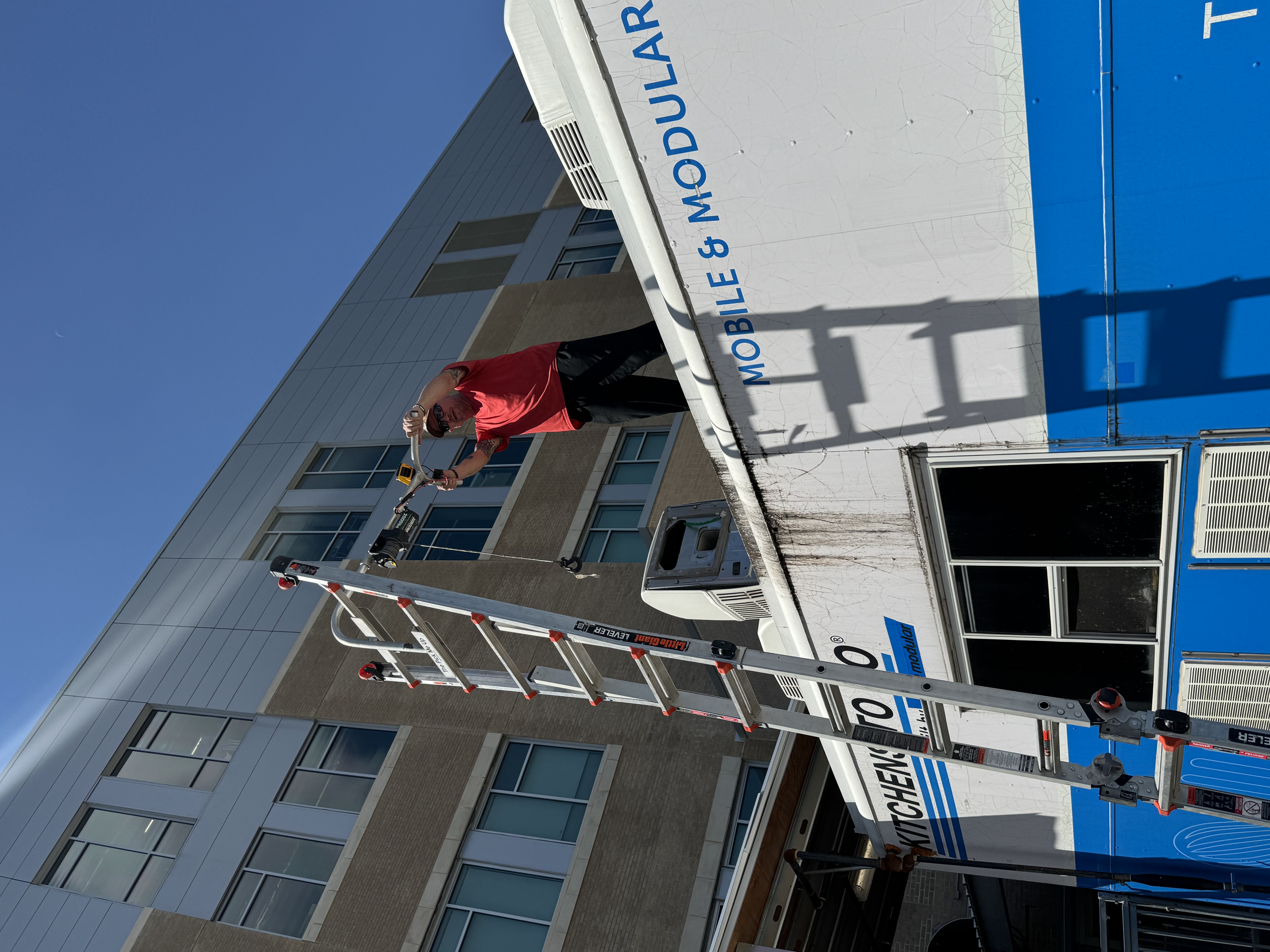 Boss Bull technician standing on the roof of a work trailer using a hoist system to lower an RV air conditioner unit during an AC replacement service call — Boss Bull Mobile RV Services