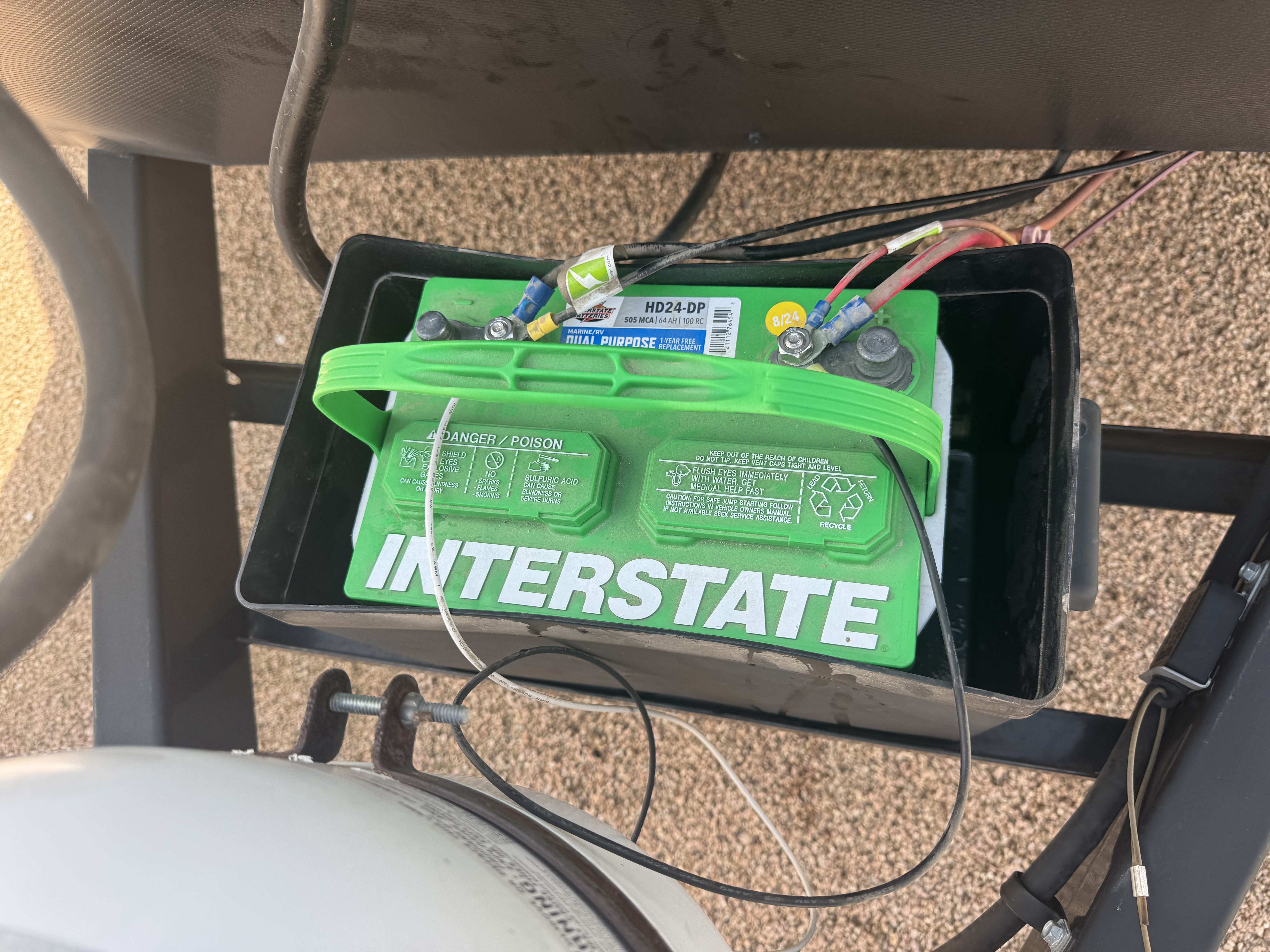 Top-down view of a black RV battery tray with a new green Interstate HD24-DP 12V battery installed, blue ring terminal connections on both posts, LP tank visible to the left — battery replacement service — Boss Bull Mobile RV Services