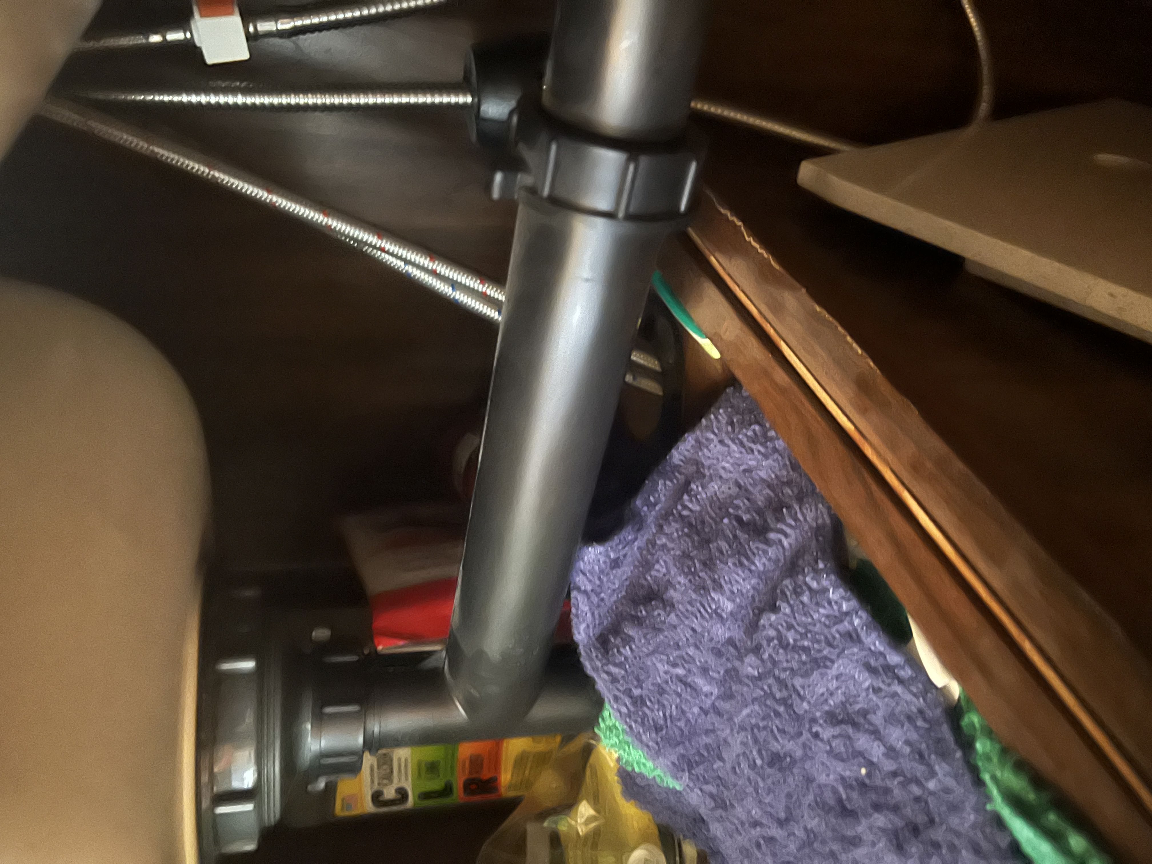 Under-cabinet view of an RV kitchen sink showing PVC P-trap, slip-joint drain fittings, and braided stainless supply lines with a towel catching an active drip — Boss Bull Mobile RV Services