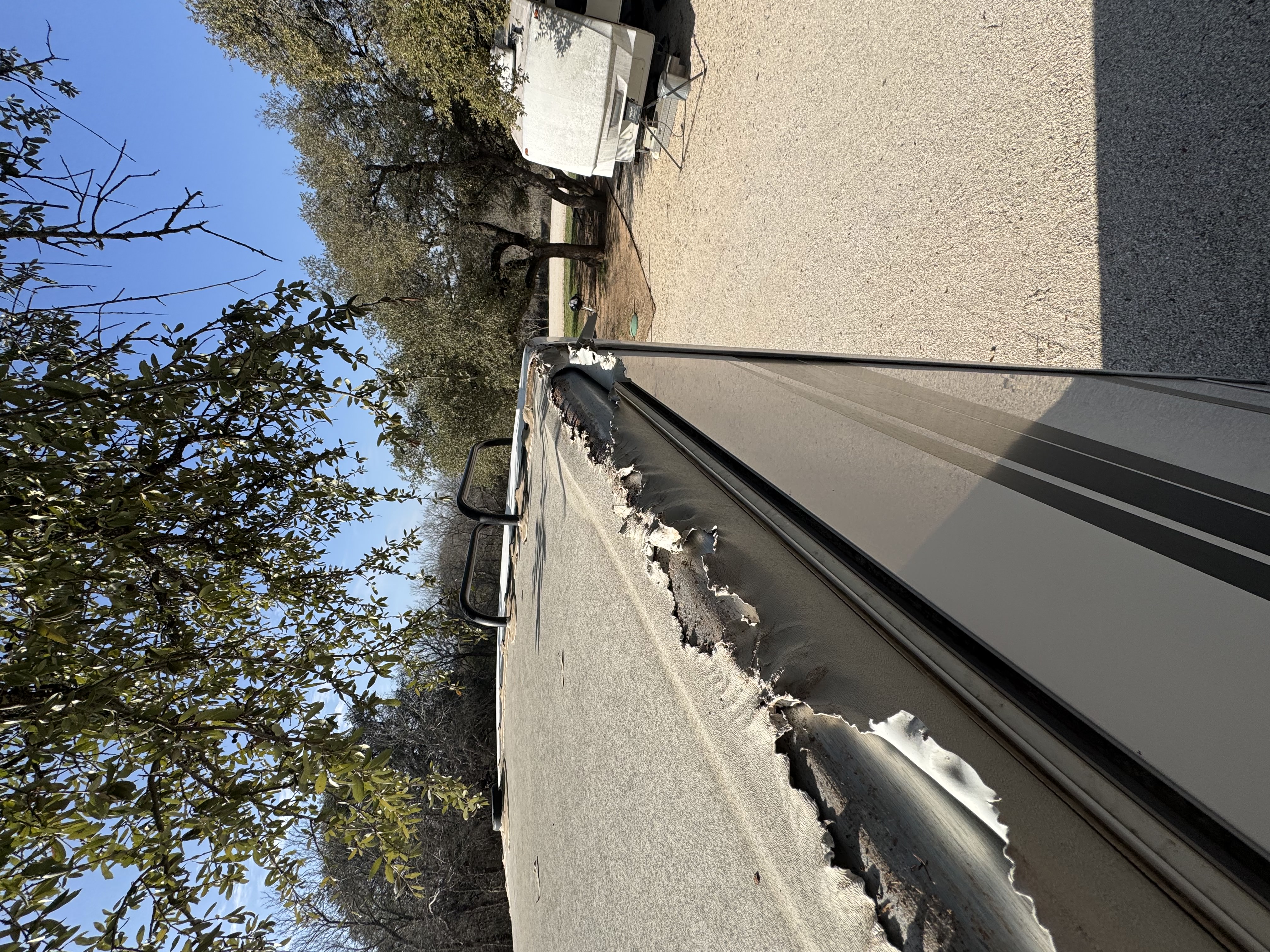 View from above looking down the drip edge of an RV at a campsite — rubber roof membrane peeling back from the aluminum rail in a ragged strip — delamination documented before repair — Boss Bull Mobile RV Services