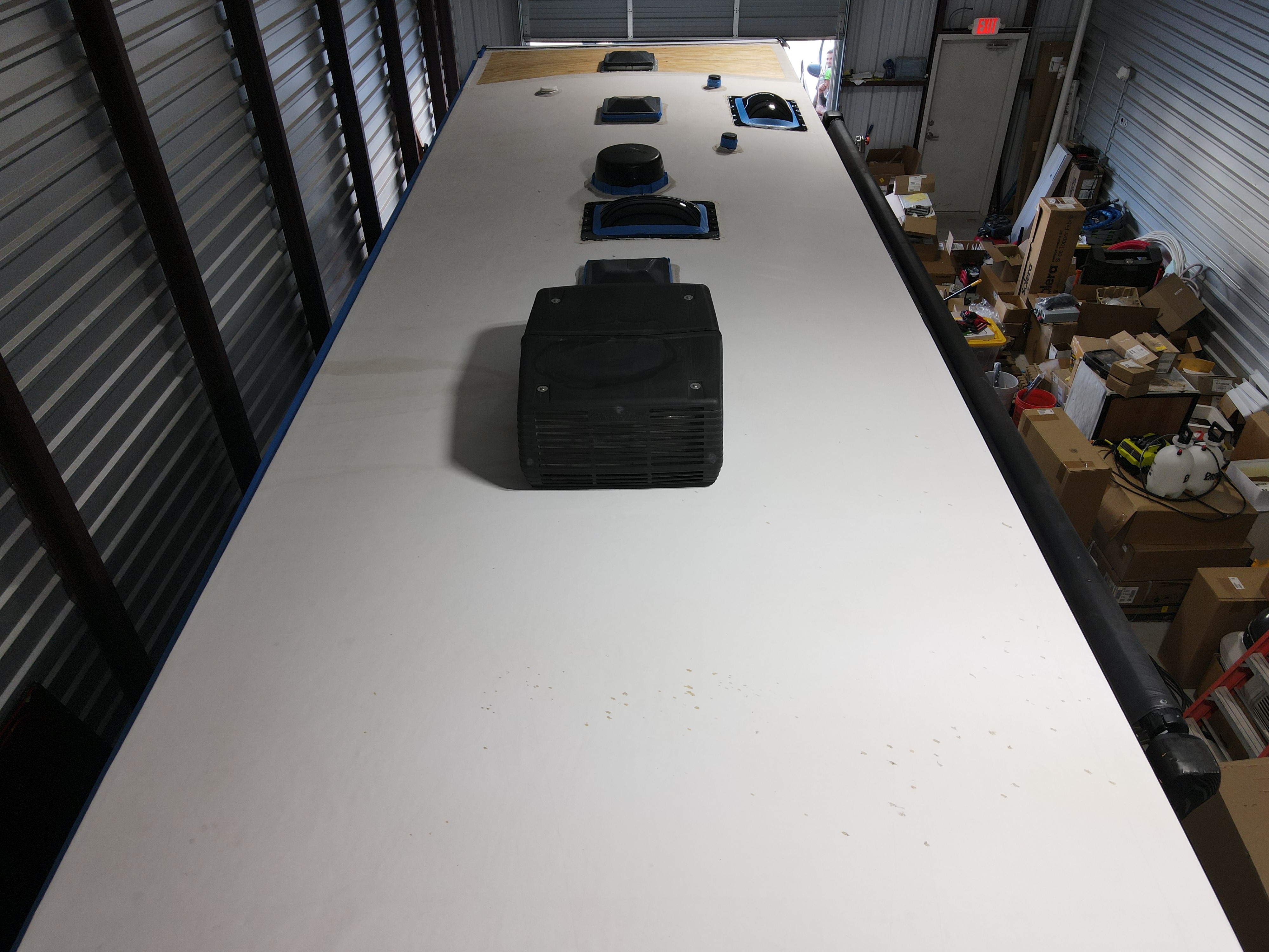 Shop interior view looking forward from the rear of an RV roof — new white rubber membrane fully laid and all penetrations blue-tape masked before elastomeric coating — six penetrations visible including AC unit, dome vent, skylights, and satellite dome — Boss Bull Mobile RV Services