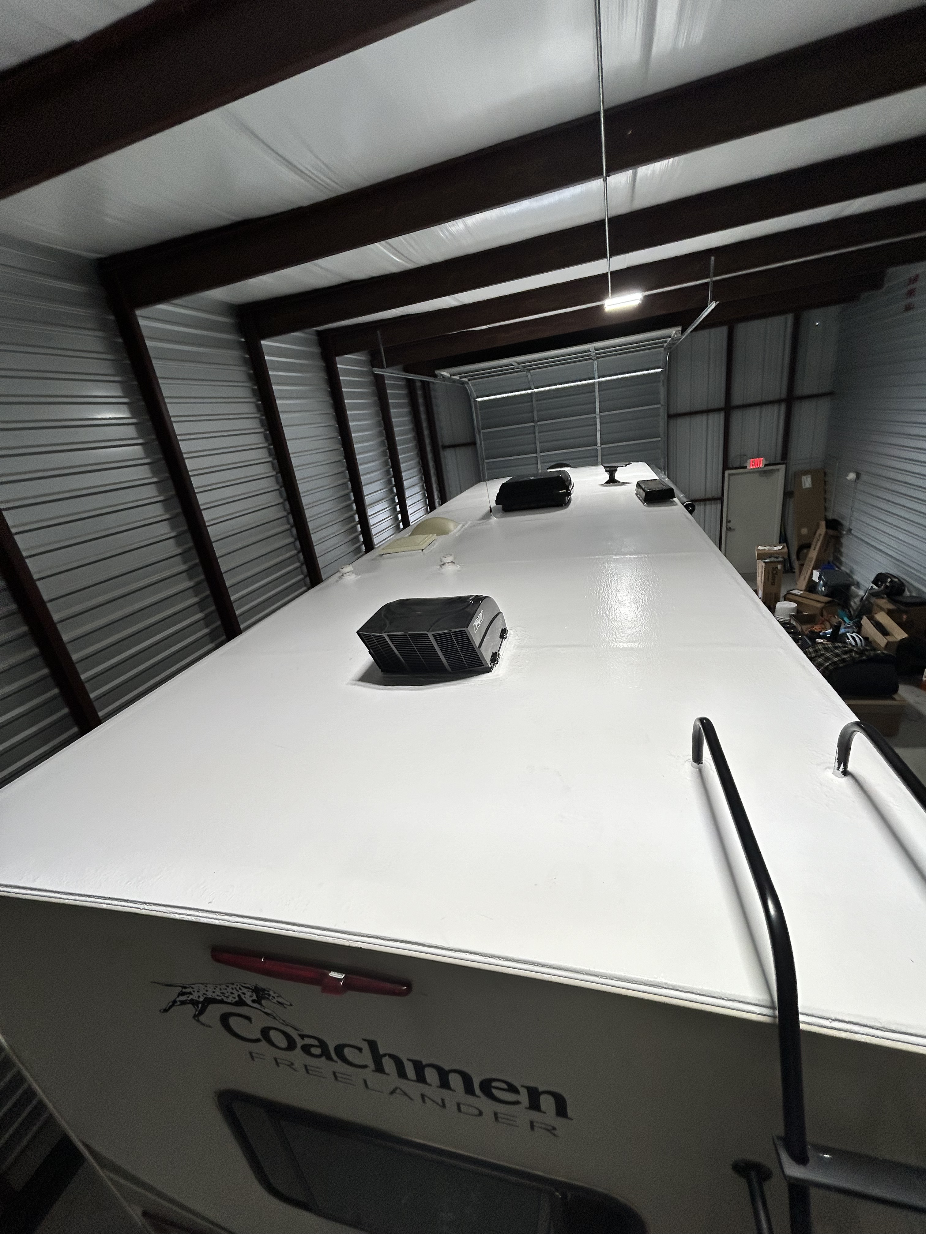 Wide-angle shop view of a Coachmen Freelander RV during mid-stage roof restoration — approximately 80 percent of the roof surface white-coated, one skylight masked — Boss Bull Mobile RV Services