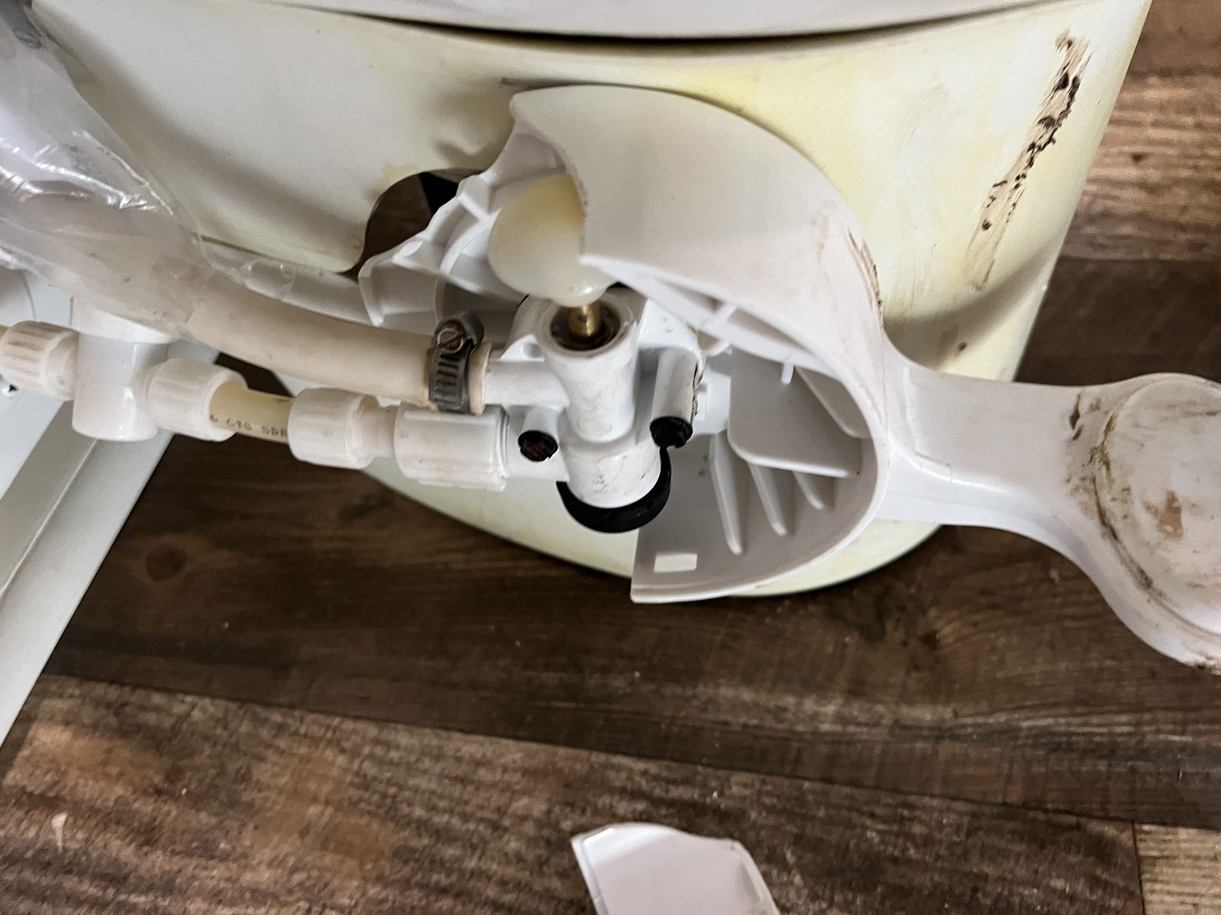 RV toilet tipped on its side showing the underside with white PVC manifold, brass supply fitting, hose clamp, and water valve components — failed valve documented for repair — Boss Bull Mobile RV Services