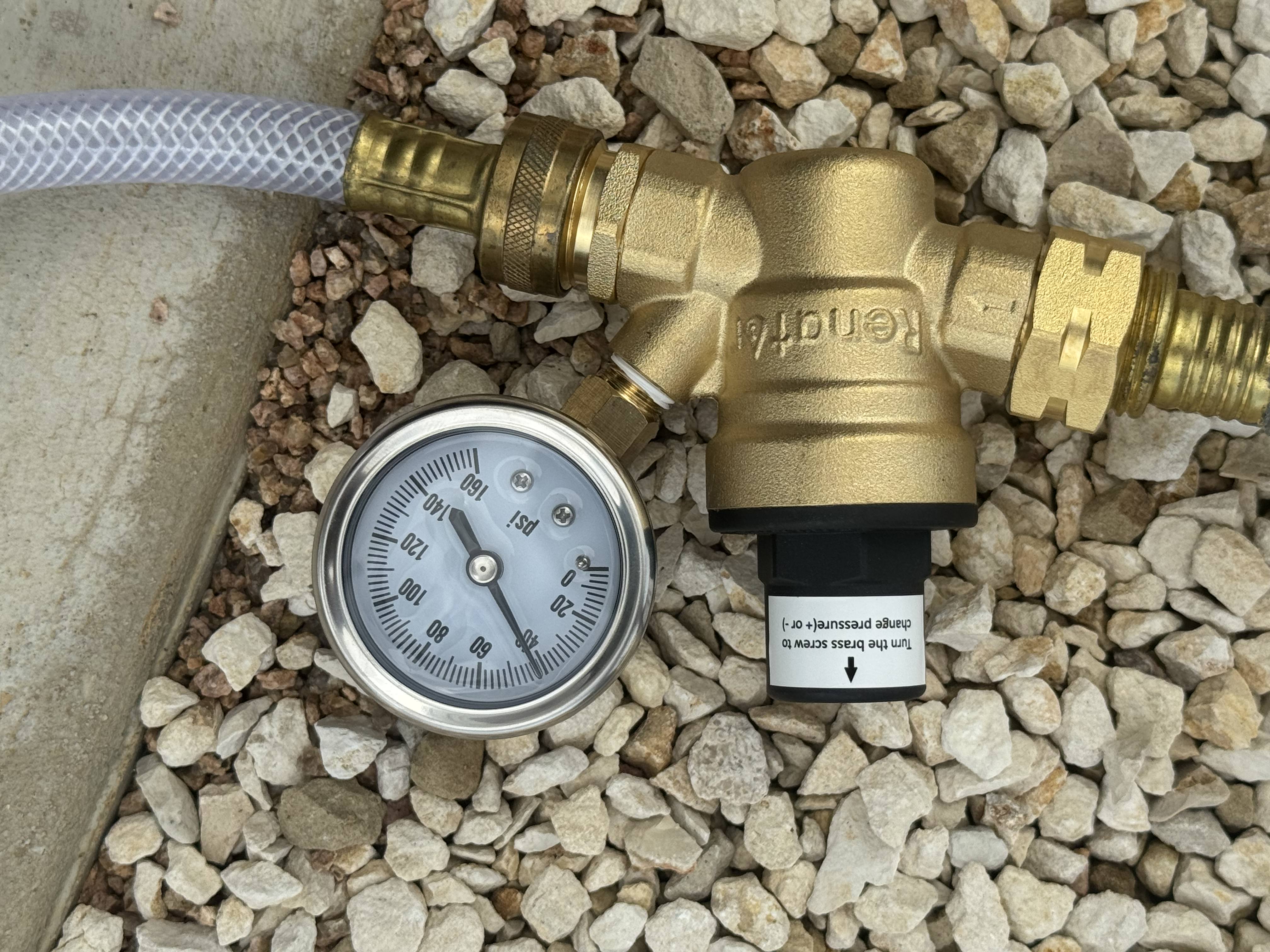 Brass Renator RV water pressure regulator with inline PSI gauge reading approximately 40 PSI — installed and tested with a white braided water hose on a gravel surface — Boss Bull Mobile RV Services