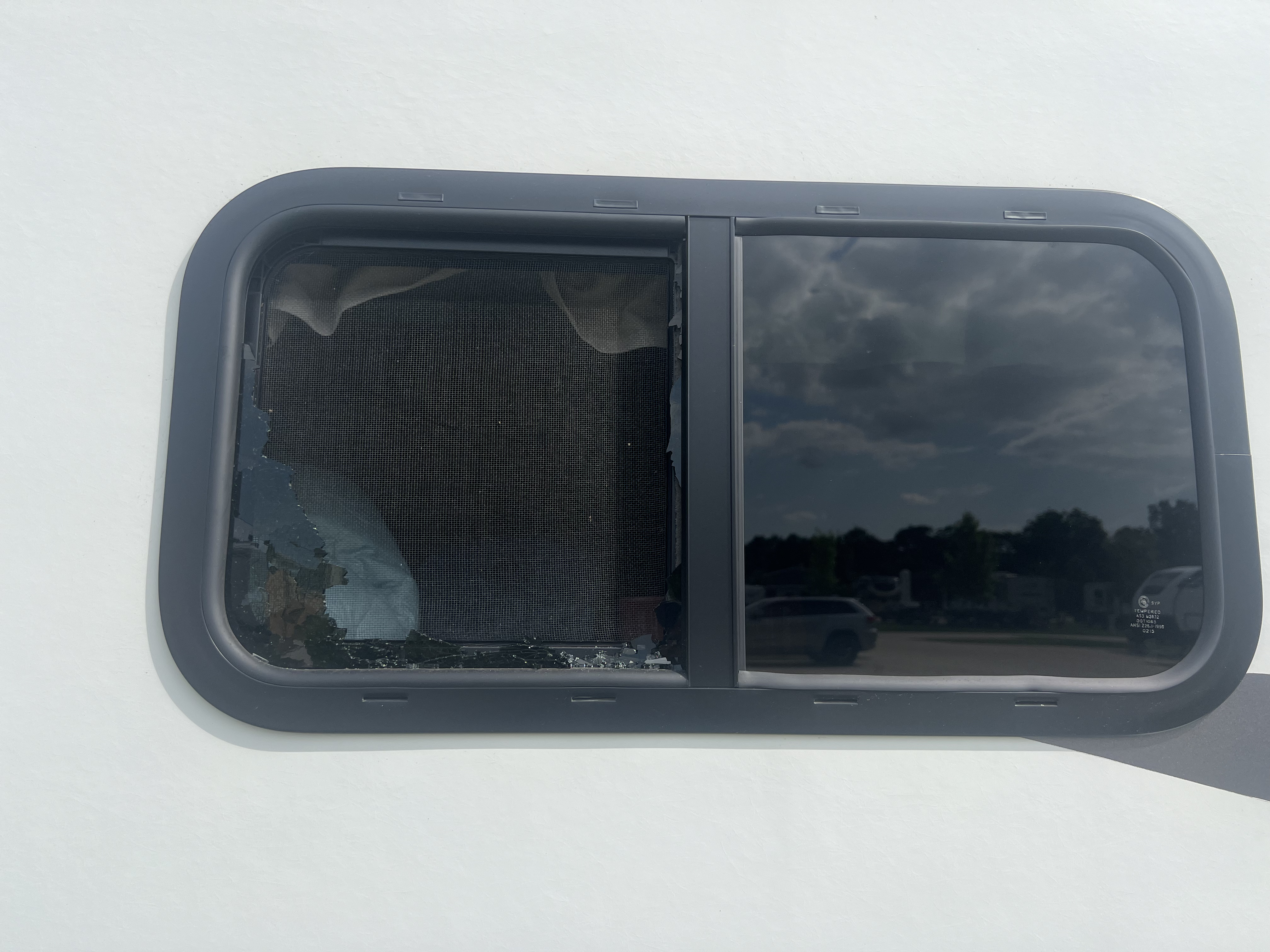 White-sided RV exterior showing a dual-pane slider window with the left pane fully shattered — glass punched through and screen torn away, right pane fully intact — damage documented before insurance claim — Boss Bull Mobile RV Services
